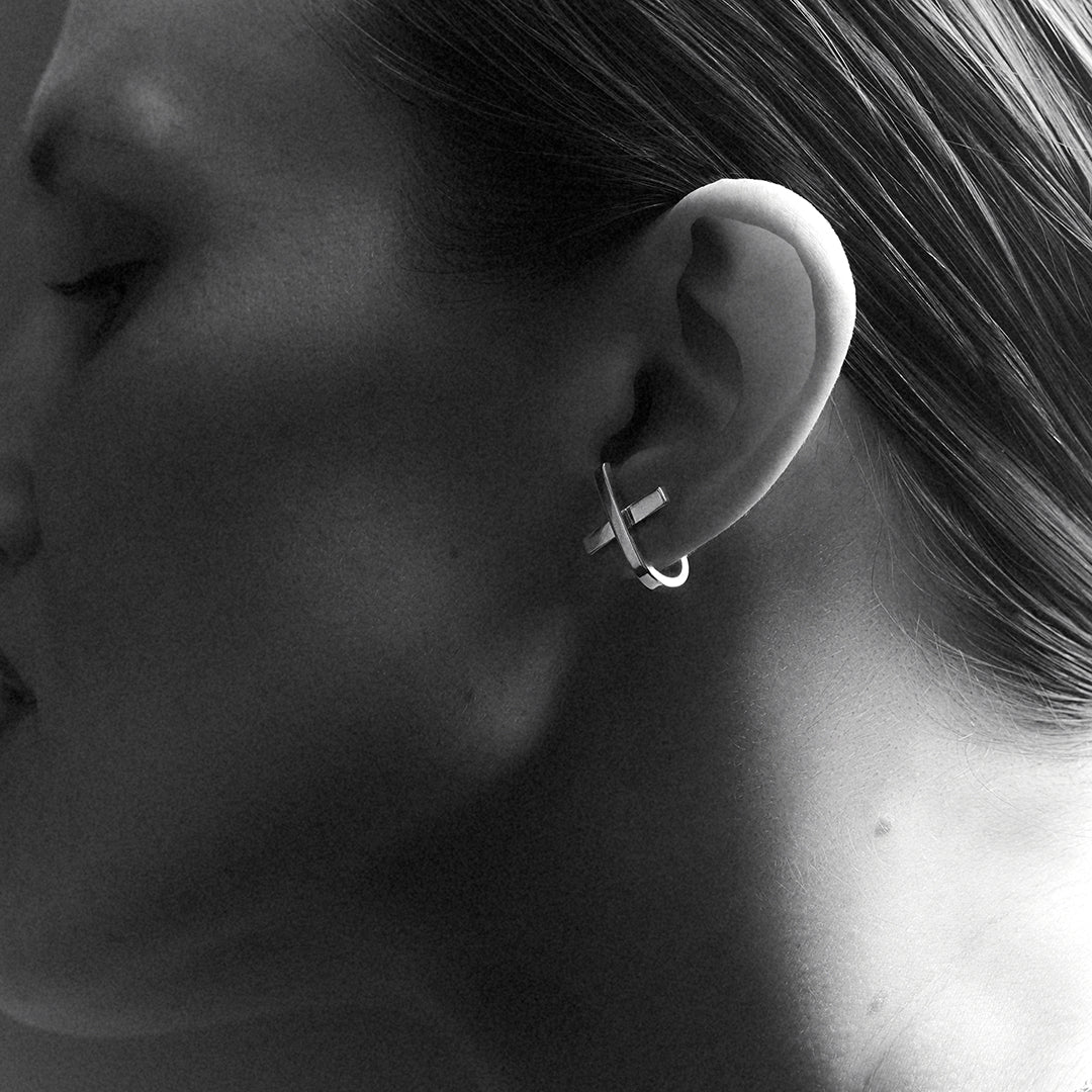 A woman's profile wearing an x or cross shaped silver earring with a hoop that hugs around her earlobe.