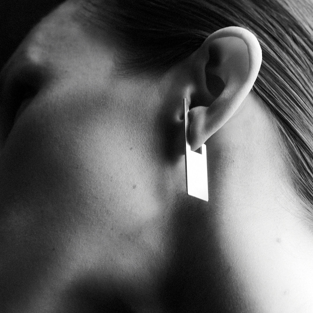 A woman's side profile zoom in, wearing a rectangular shaped earring which hoops onto her earlobe.