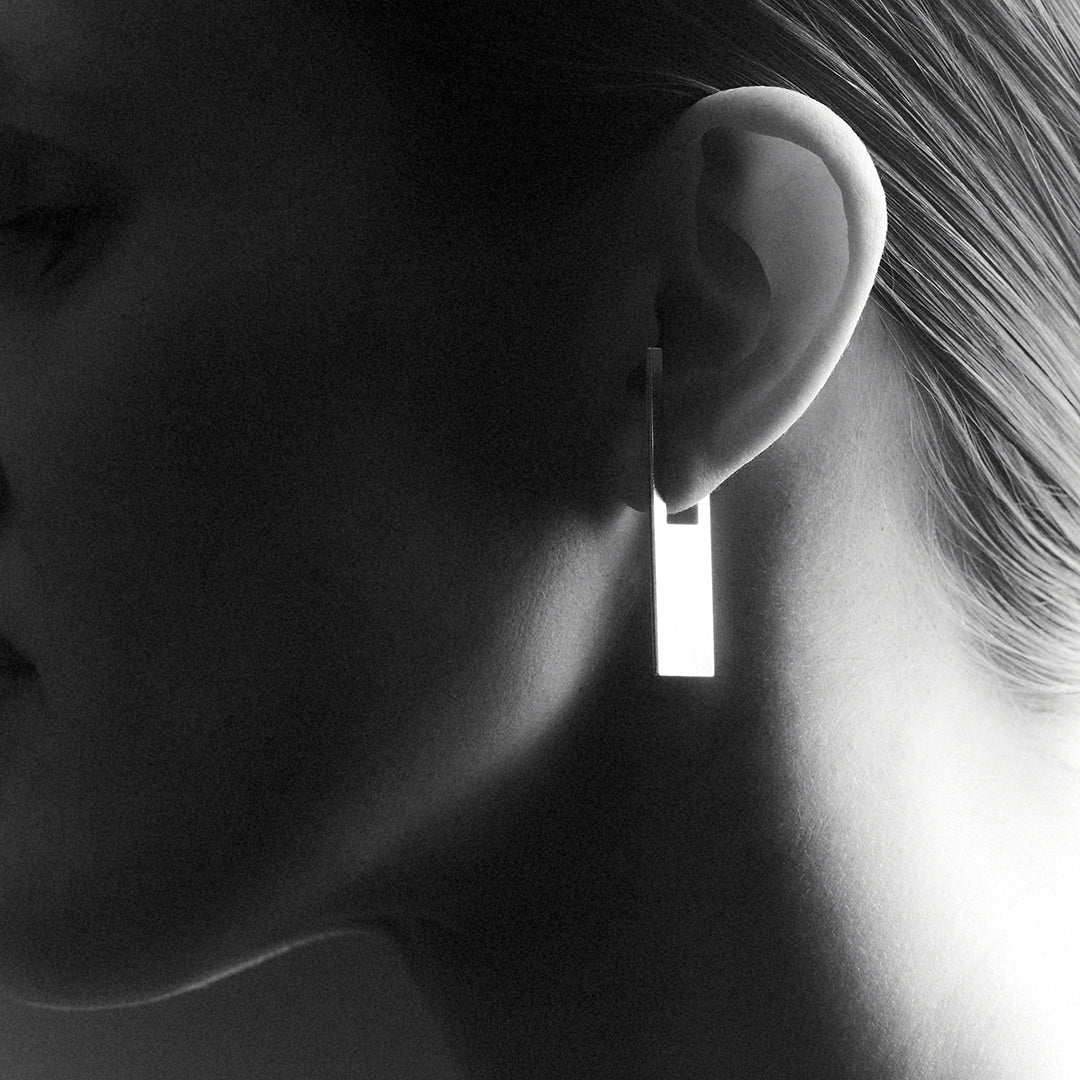 A woman's side profile zoom in, wearing a rectangular shaped earring which hoops onto her earlobe. 