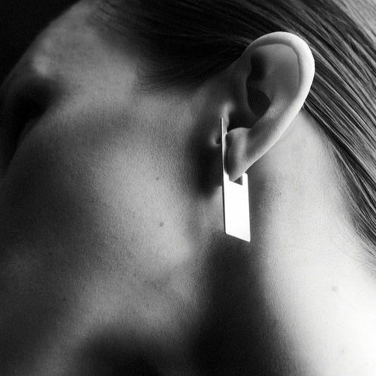 A woman's side profile zoom in, wearing a rectangular shaped earring which hoops onto her earlobe. 