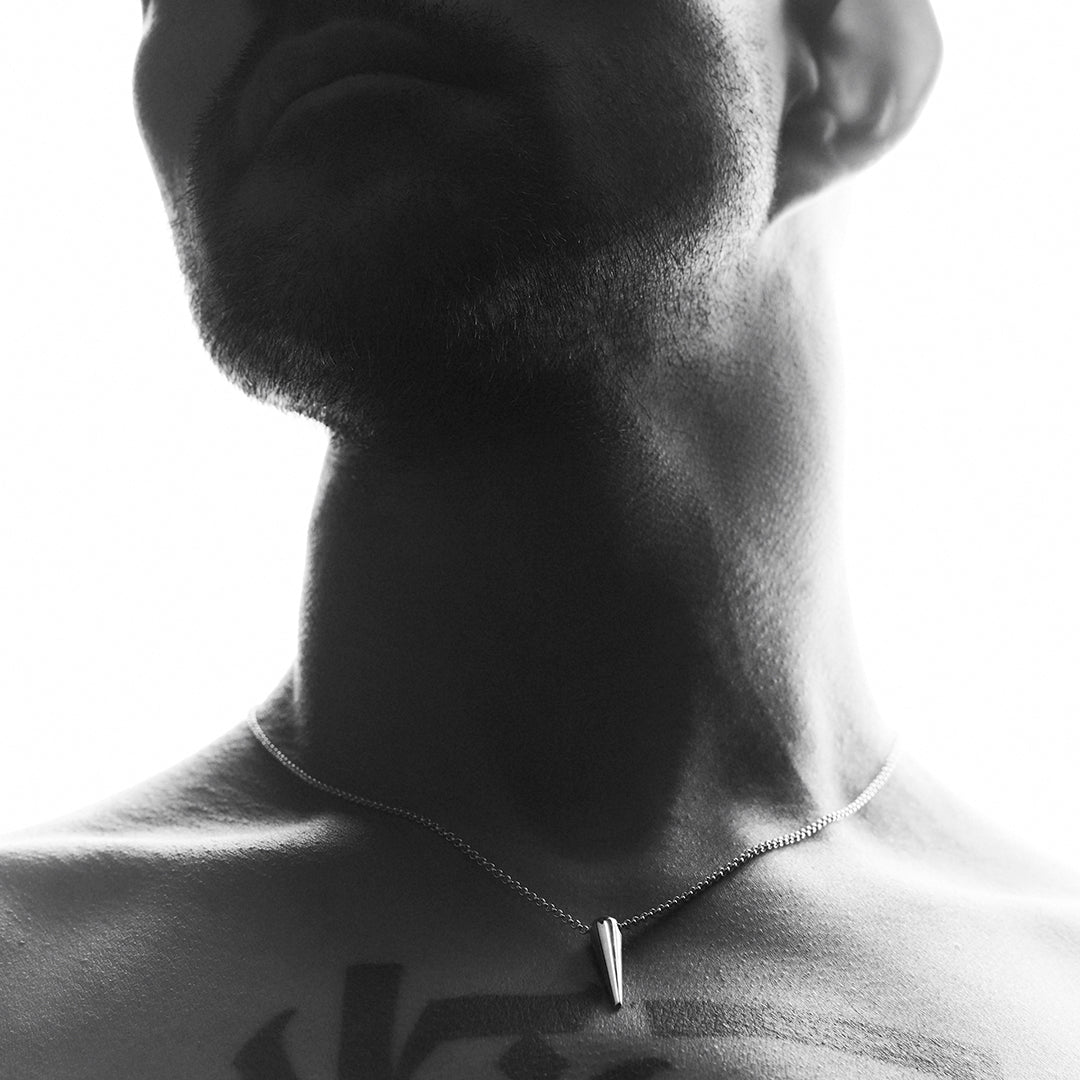 A man's chest and neck, with a silver chain and a silver spike shaped pendant. 