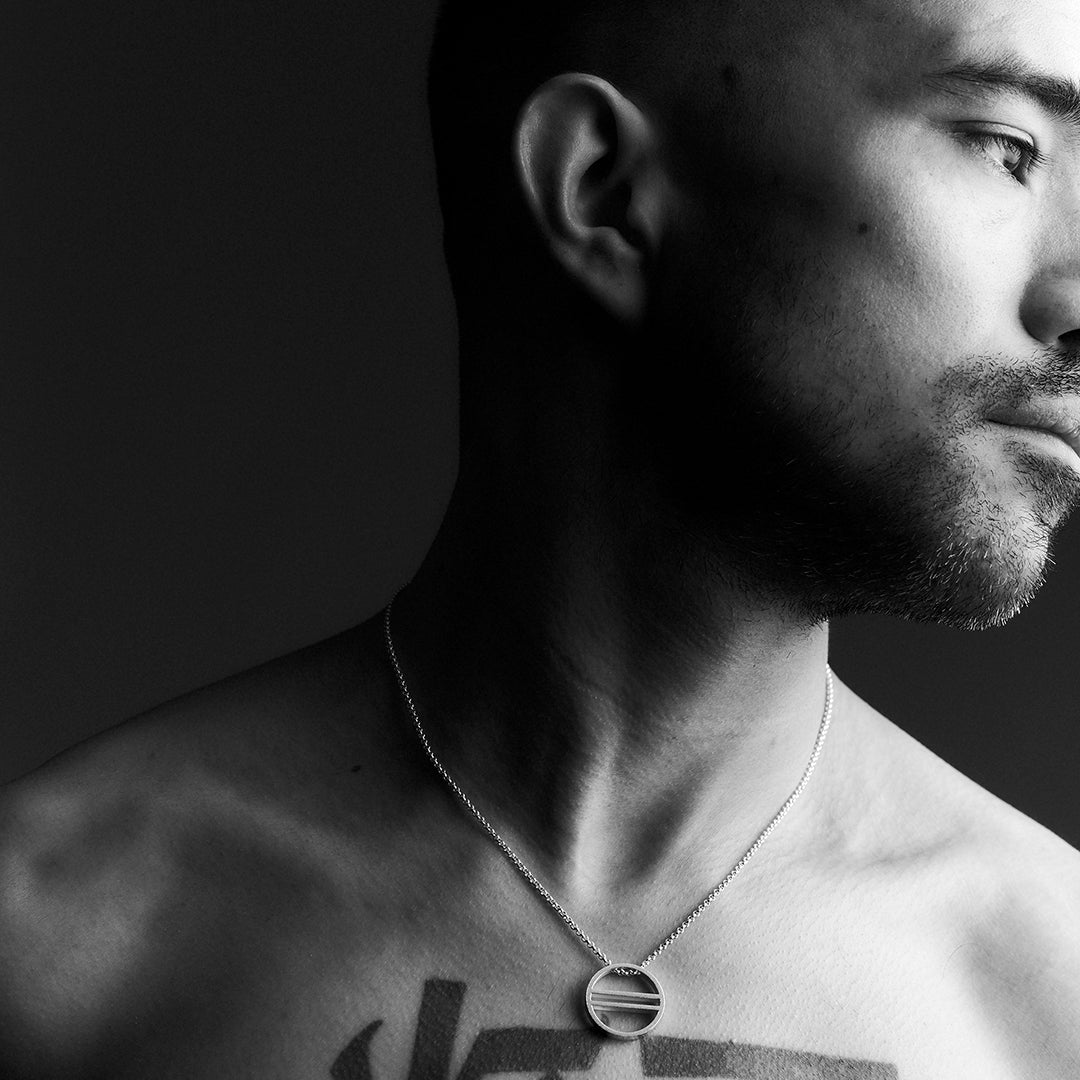 A man's face & chest wearing a silver necklace with an equality symbol pendant. 